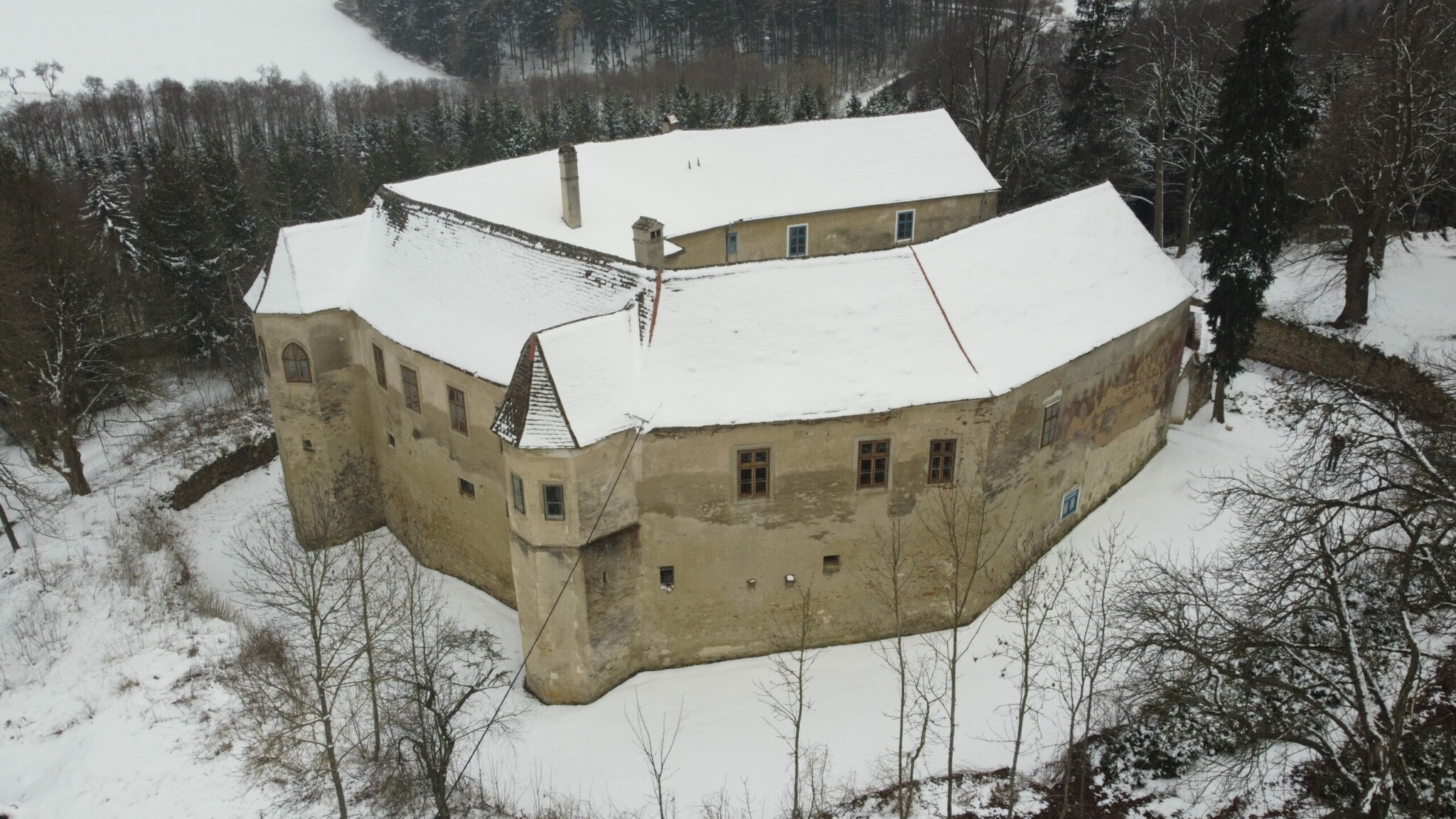 An aerial photo captures the historic, three-winged structure of Schloss Lichtenau im Waldviertel, surrounded by a defensive wall, a dry moat, and lush greenery, with a small part of the village visible beyond.